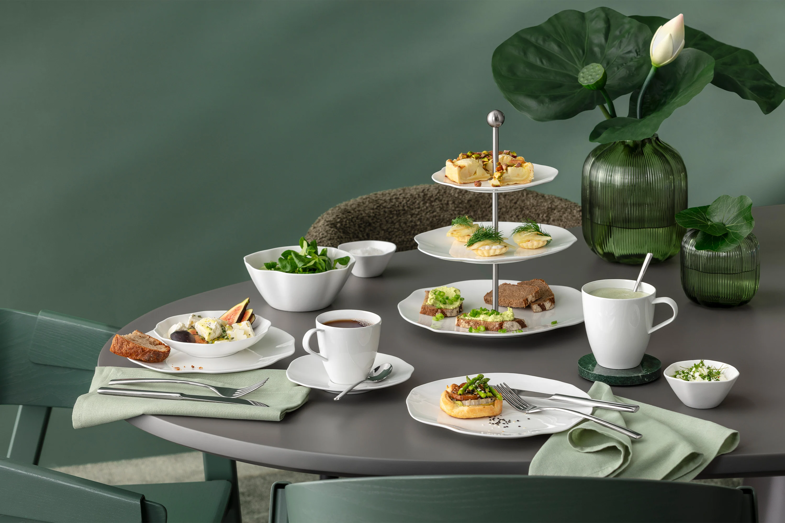 A round table with white tableware from the Pura collection, various baked goods, salad, tea and green leaf decorations against a green background.