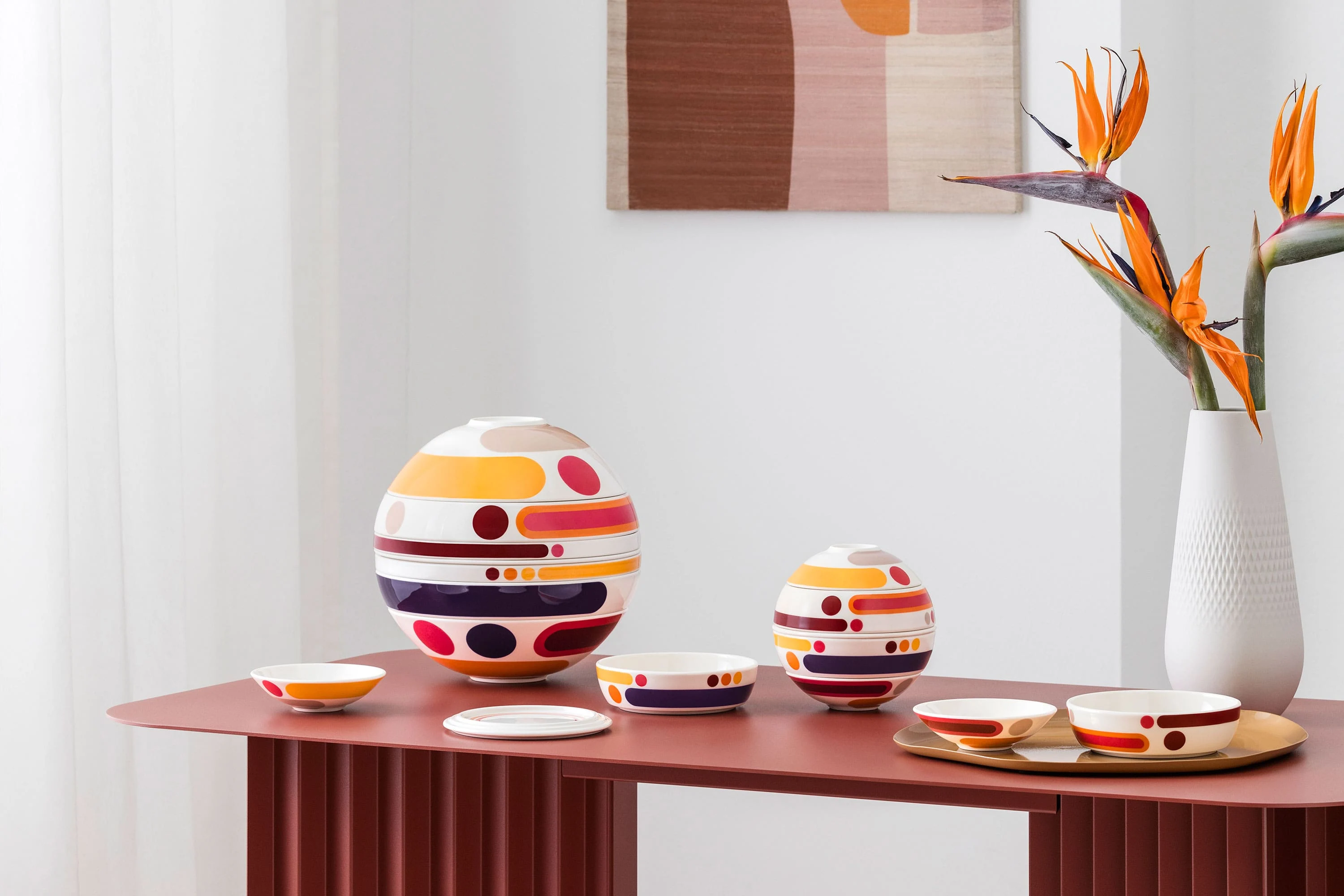 La petite boule miami and la boule miami arranged on a red table Colorful, multi-piece tableware sets from la petite boule miami, circular design objects in large and small sizes, stand as decoration next to a vase with abstract patterns and orange flowers on a red table.