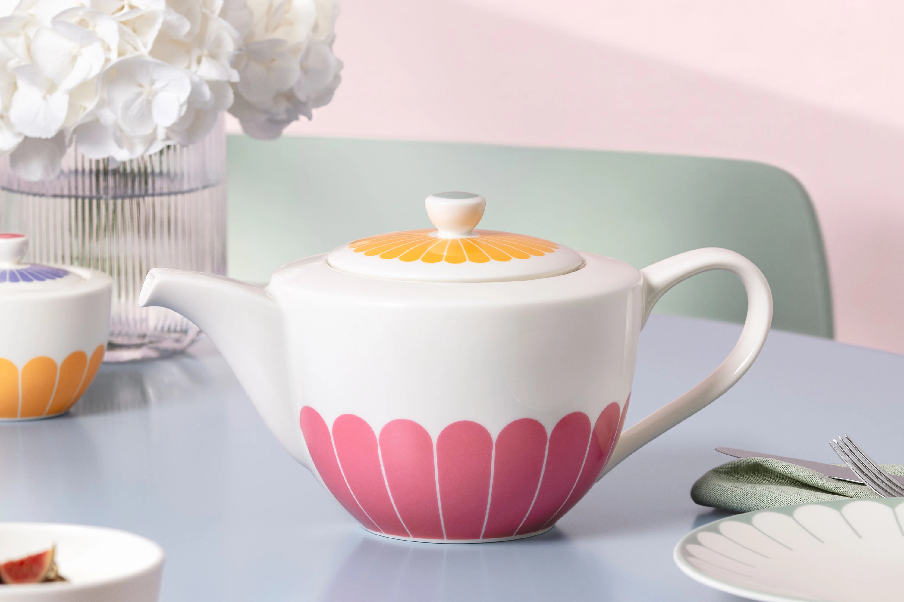 A white milk jug by Fleur with a yellow lid and pink leaf pattern stands on a light blue table next to a cup and white flowers.