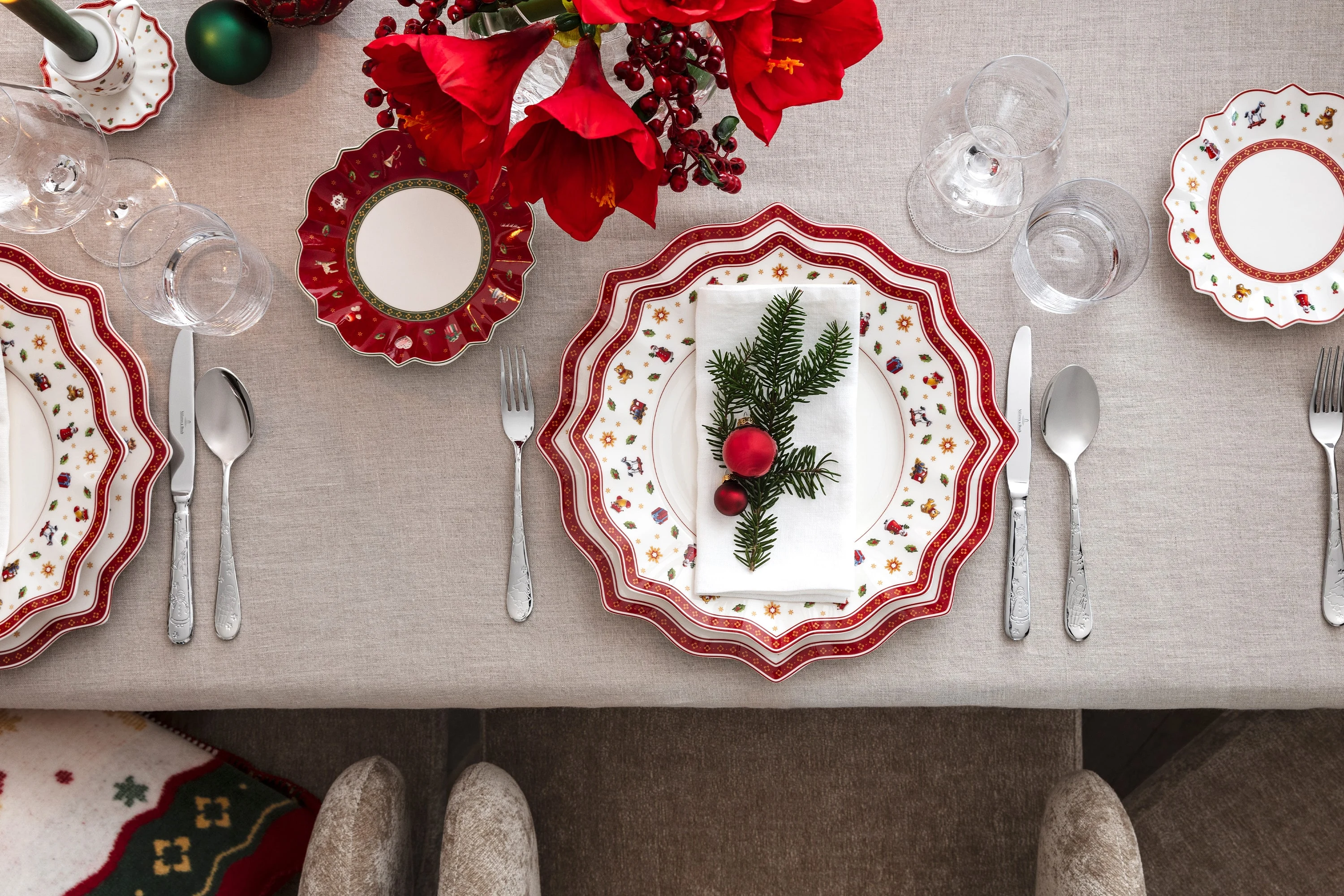 Top view of an elegant festive table with the Villeroy & Boch Toy's Delight collection, complete with red and white plates, silver cutlery and a festive napkin decorated with pine wood and ornaments.