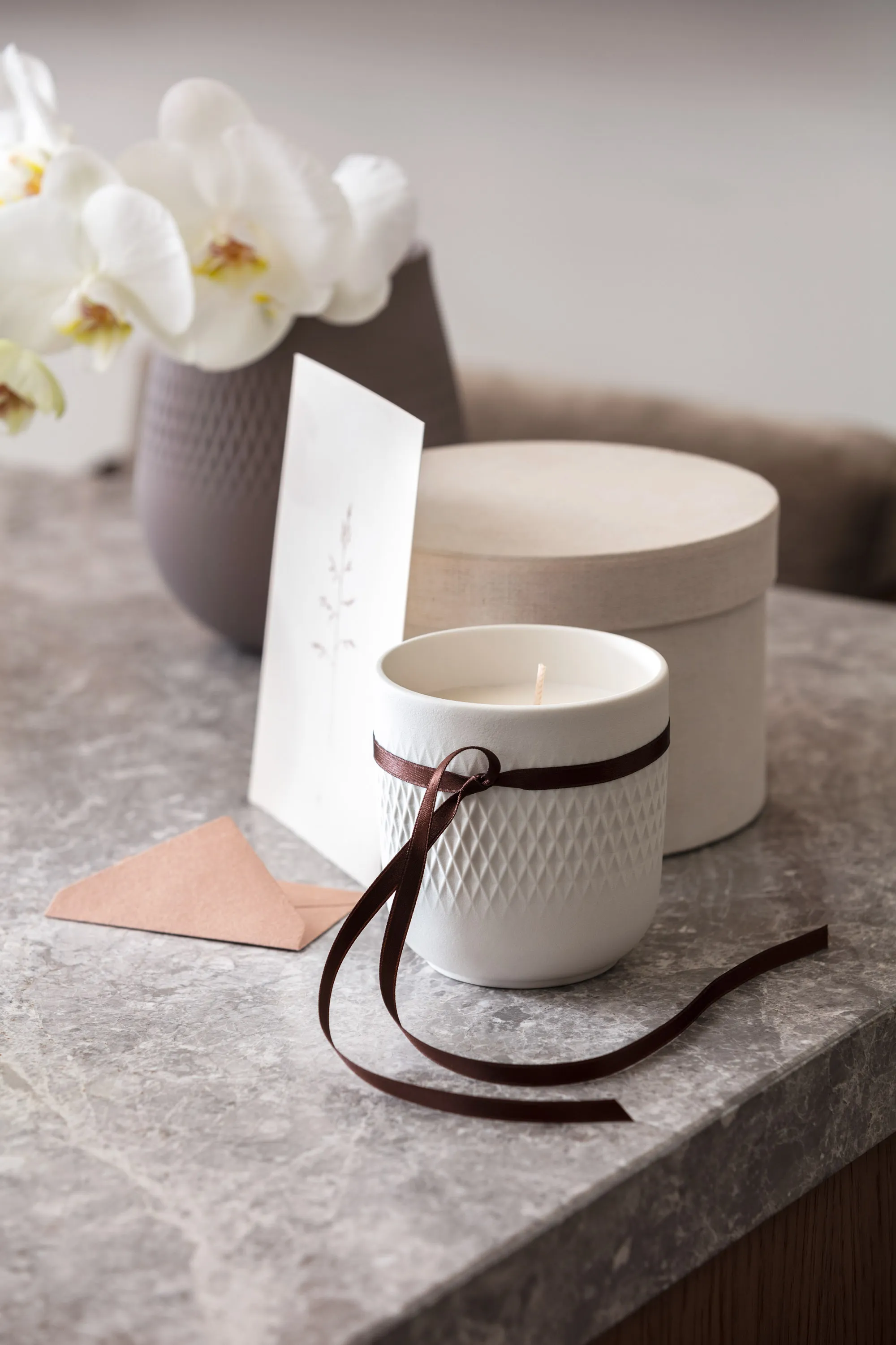 Une bougie parfumée Manufacture en blanc dans un emballage cadeau Une bougie parfumée Manufacture blanche de Villeroy & Boch emballée en cadeau avec un ruban marron et une boîte claire en arrière-plan.