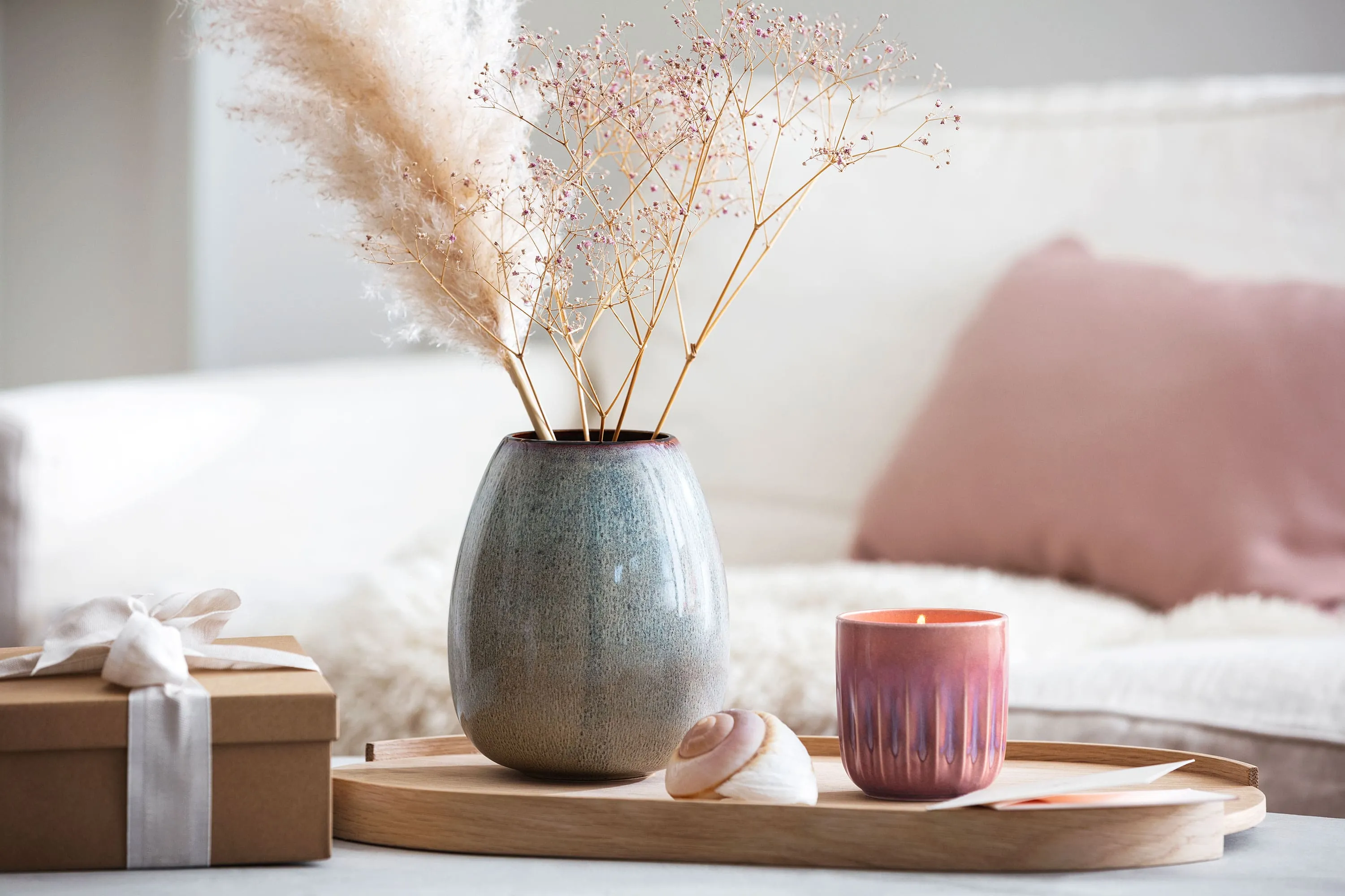 Un plateau en bois avec un vase Lave et une bougie Perlemor Un plateau en bois sur lequel sont posés un vase Lave et une bougie Perlemor avec des fleurs séchées, à côté d’un cadeau emballé.