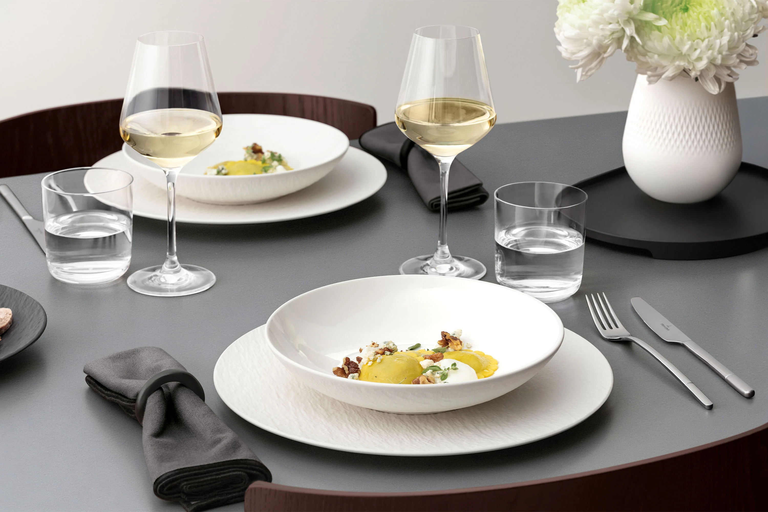 A formal table setting with a Manufacture bowl on top of a plate A formal table setting with a white bowl on a white plate, glassware, and silver cutlery on a grey table and food.