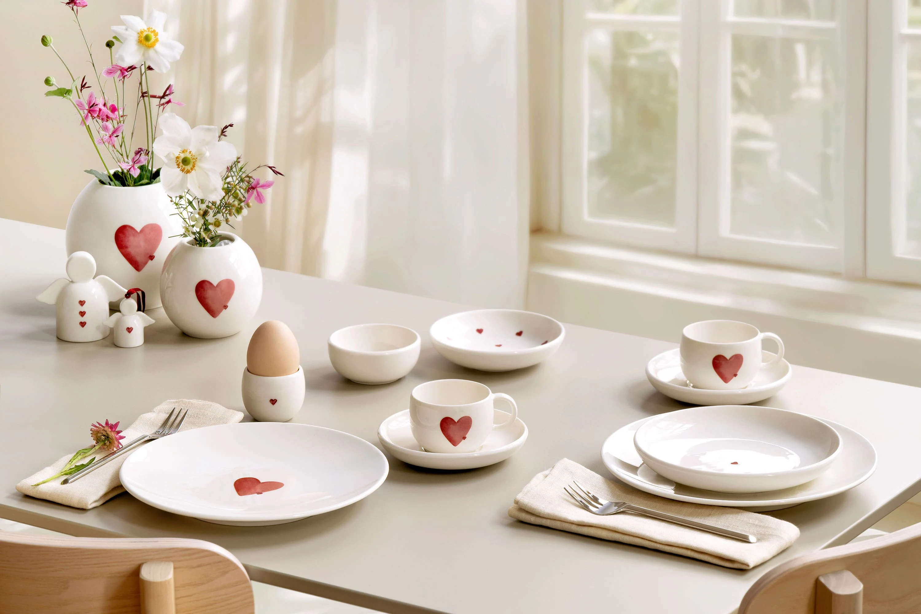 La table du petit-déjeuner est dressée avec de la vaisselle blanche ornée de cœurs rouges de with love, des fleurs dans des vases de with love et des ustensiles soigneusement disposés.