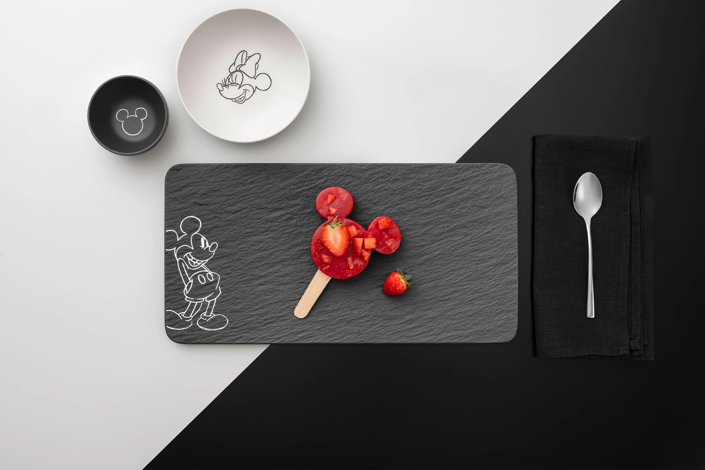 A grey Manufacture Mickey serving plate with a strawberry ice cream on a stick A grey Manufacture Mickey serving plate with a strawberry ice cream on a stick, a white bowl with seeds, a black cup and a spoon on a black napkin.