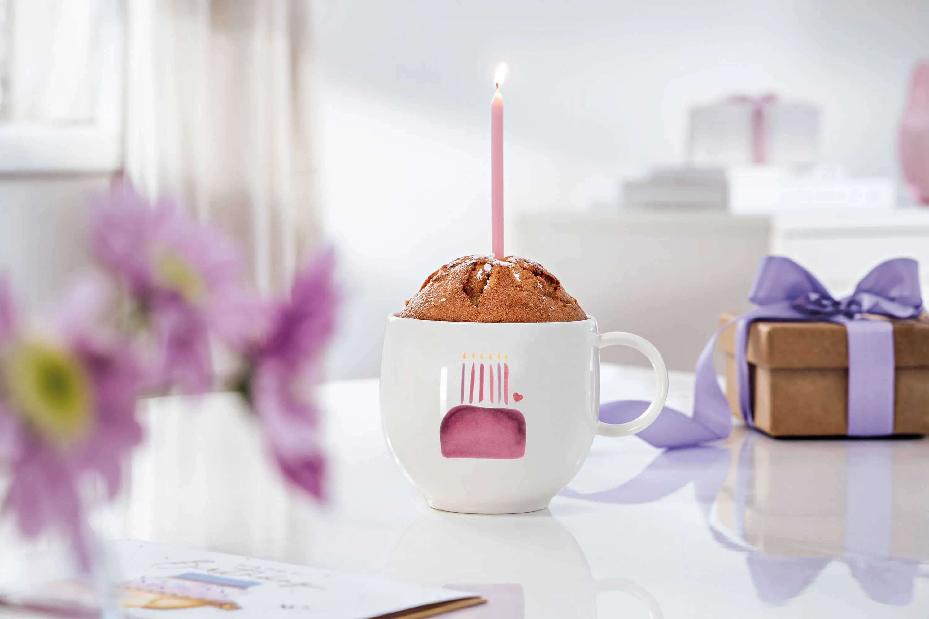 Un mugcake avec des bougies dans une tasse With Love Birthday sur une table Un mugcake avec des bougies dans une tasse With Love Birthday sur une table avec des fleurs violettes et une tasse dans une boîte cadeau beige.