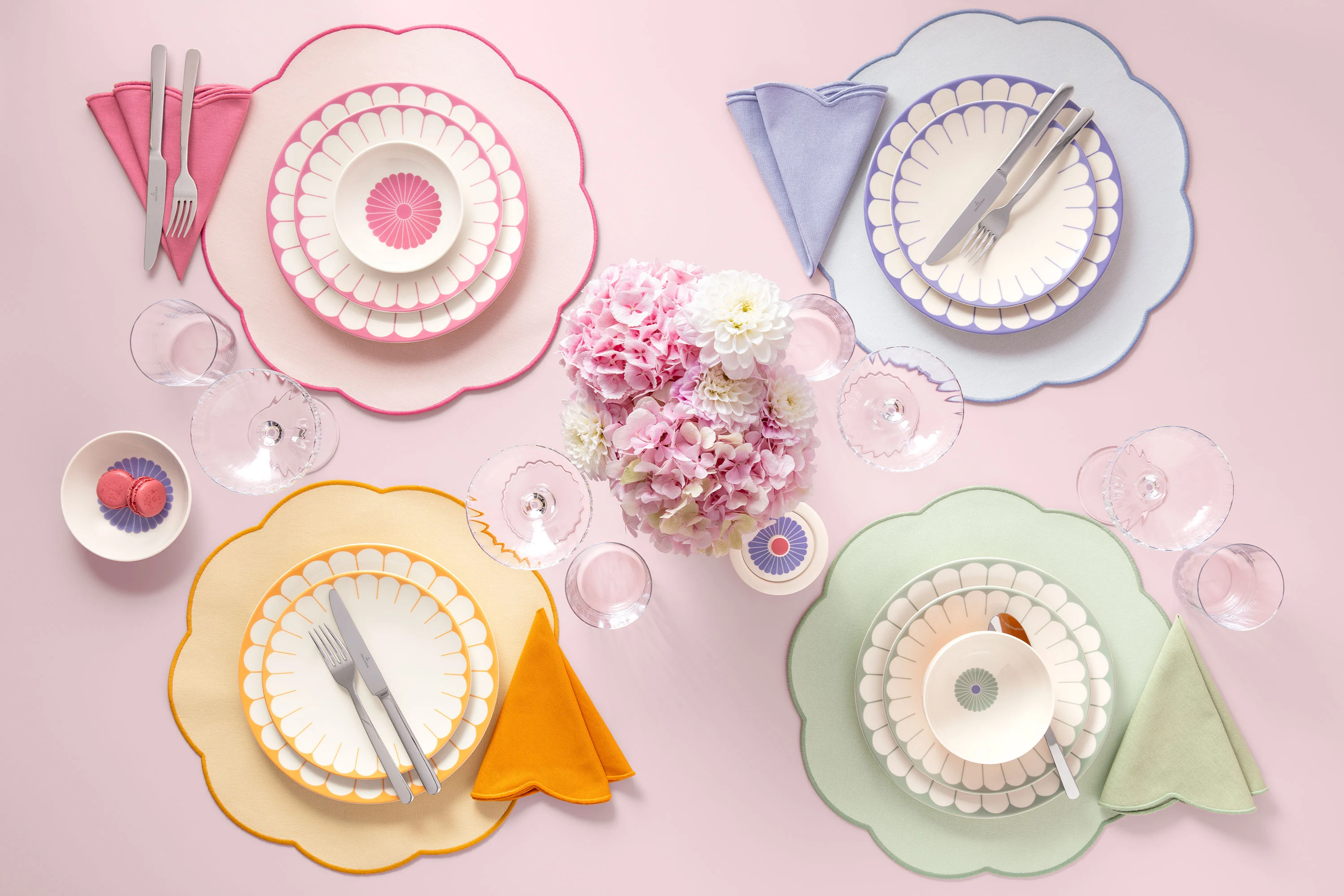 Vier pastellfarbene Gedecke der Fleur Kollektion mit geschwungenen Tellern, Servietten und Besteck umgeben einen Strauß rosa Blumen.
