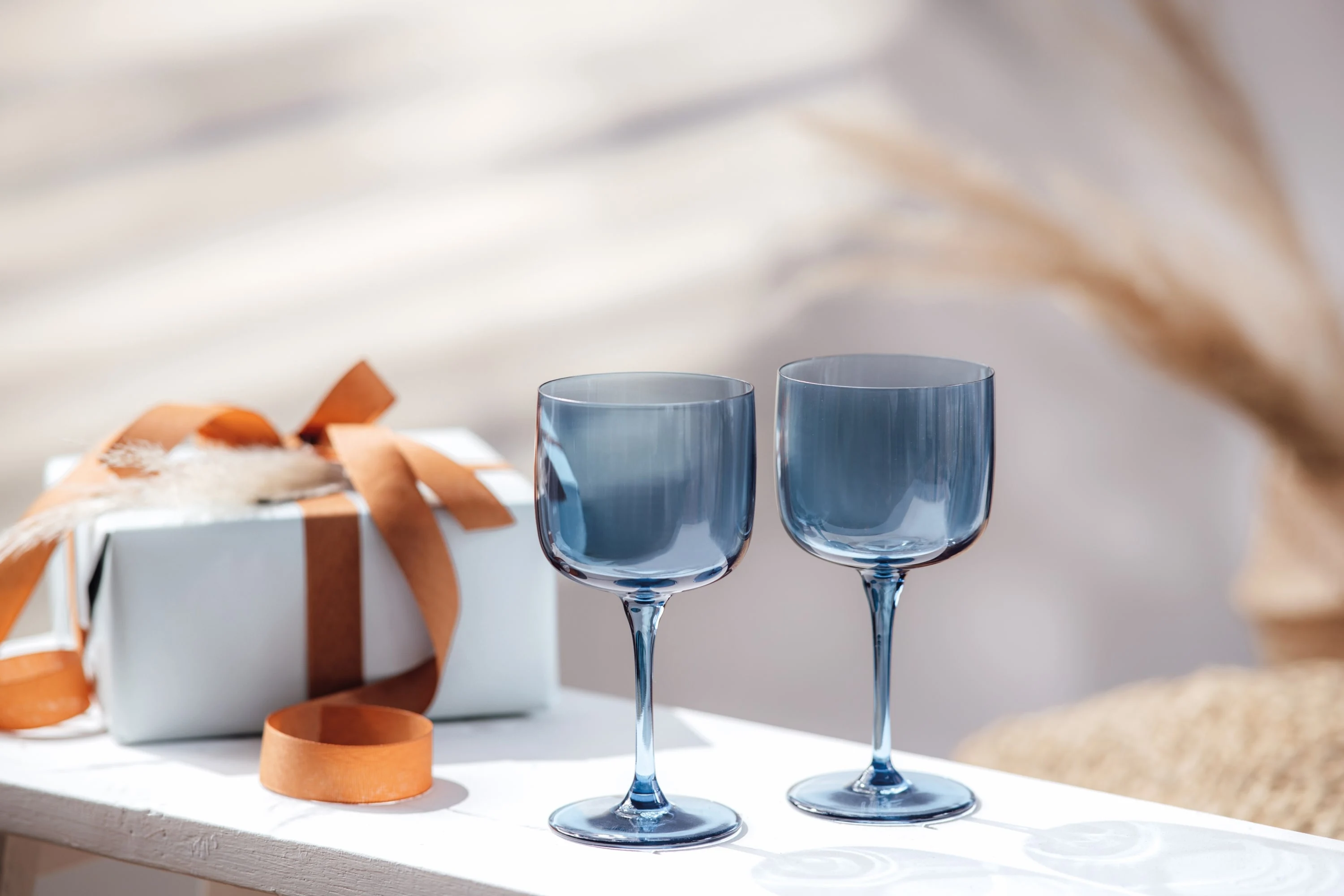 Two Villeroy & Boch Like Glass wine glasses in blue on a white table next to a wrapped gift with a brown bow.