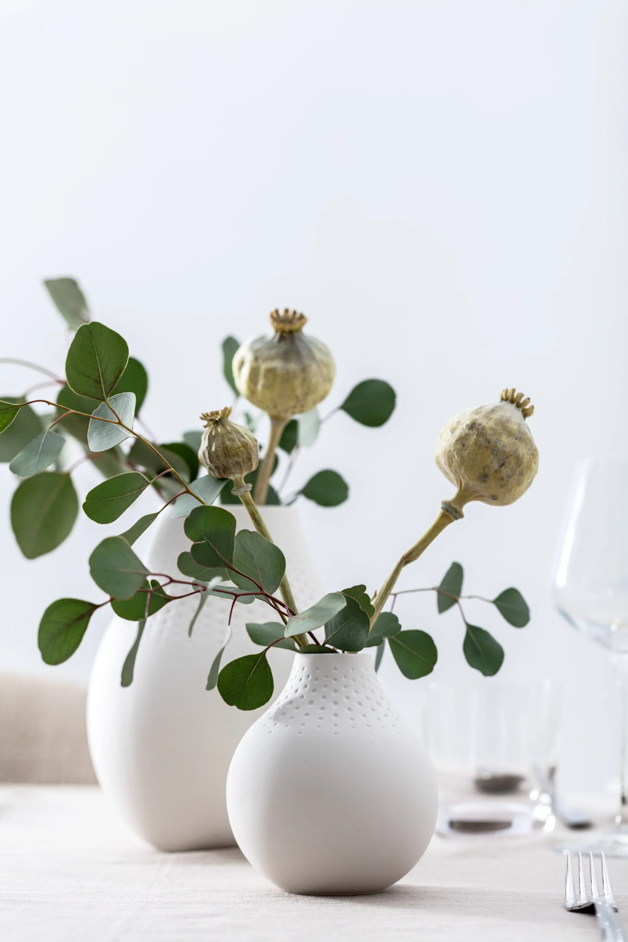 Des vases Manufacture en blanc avec de l’eucalyptus Une décoration de table minimaliste avec une branche d’eucalyptus et des capsules de pavot dans des vases blancs Manufacture.