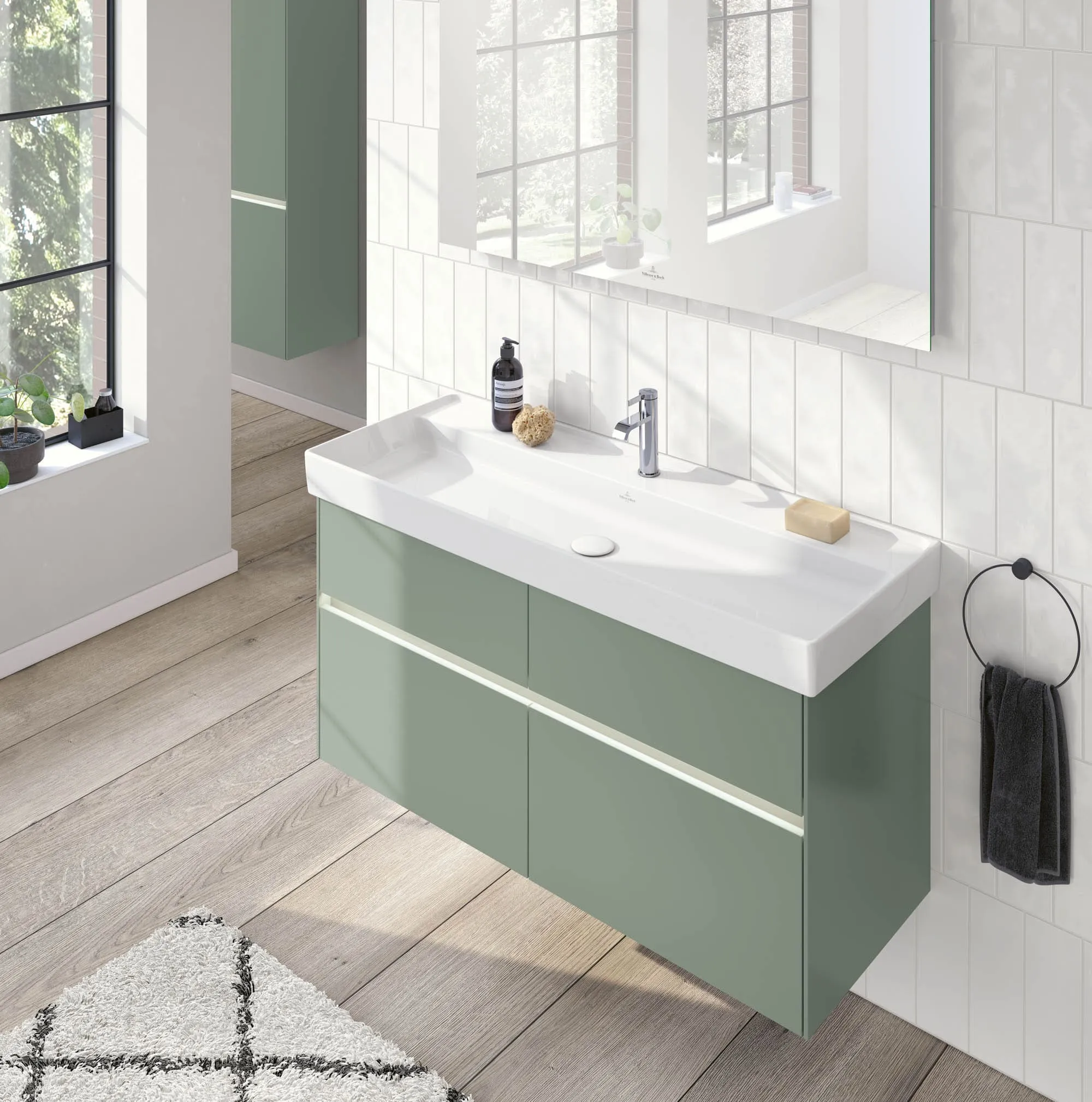 Un bagno moderno con un grande specchio, mobile lavabo verde con lavabo bianco, sapone e flacone sulla mensola. Un asciugamano pende da un portasciugamano ad anello nero. La luce naturale penetra da una finestra.