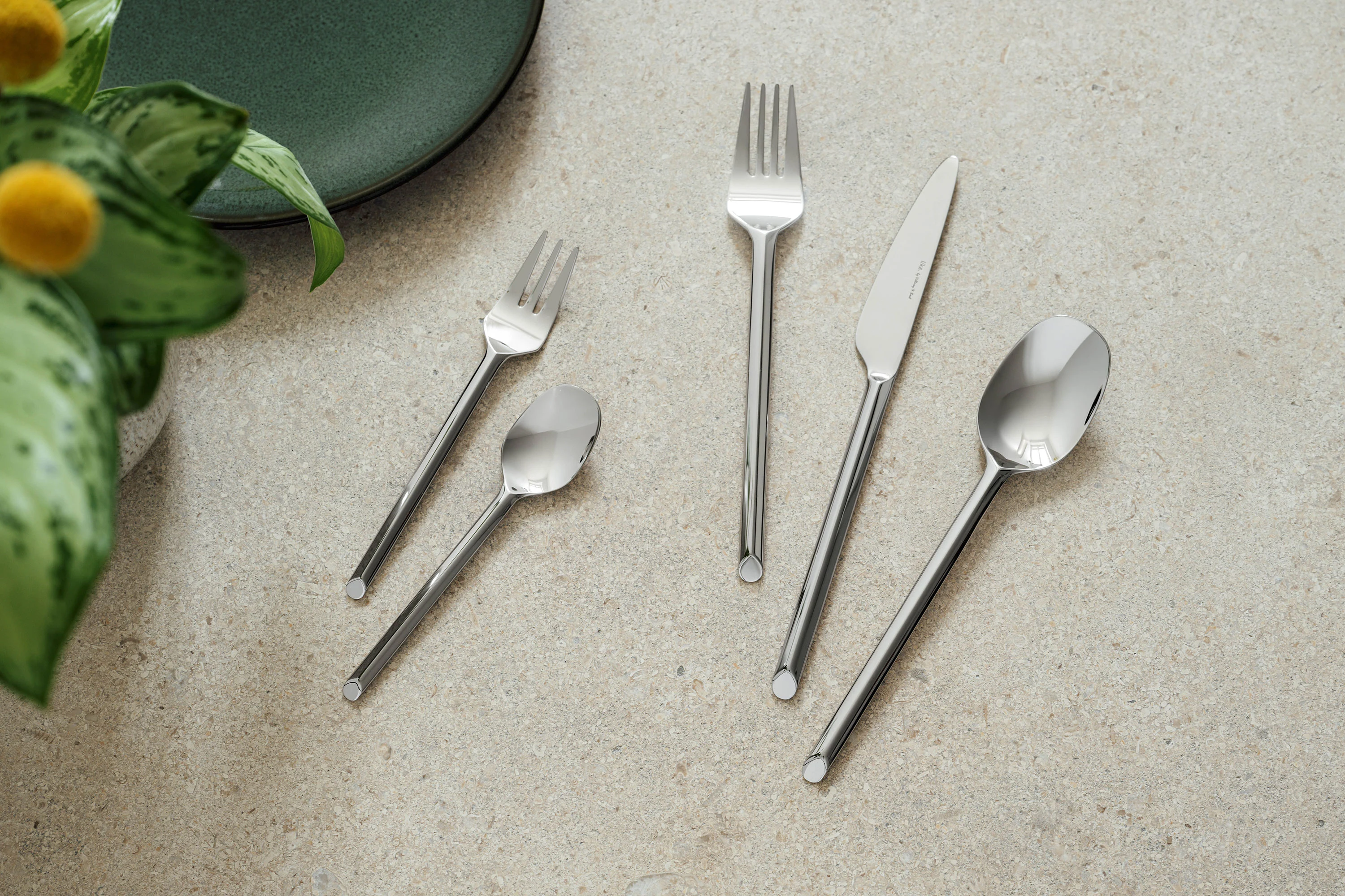 Five like cutlery pieces from like.by Villeroy & Boch- fork, knife, spoon, dessert fork and teaspoon Five pieces of cutlery in Villeroy & Boch style - fork, knife, spoon, dessert fork and teaspoon - are arranged on a light-coloured stone surface next to a green plate and a plant.