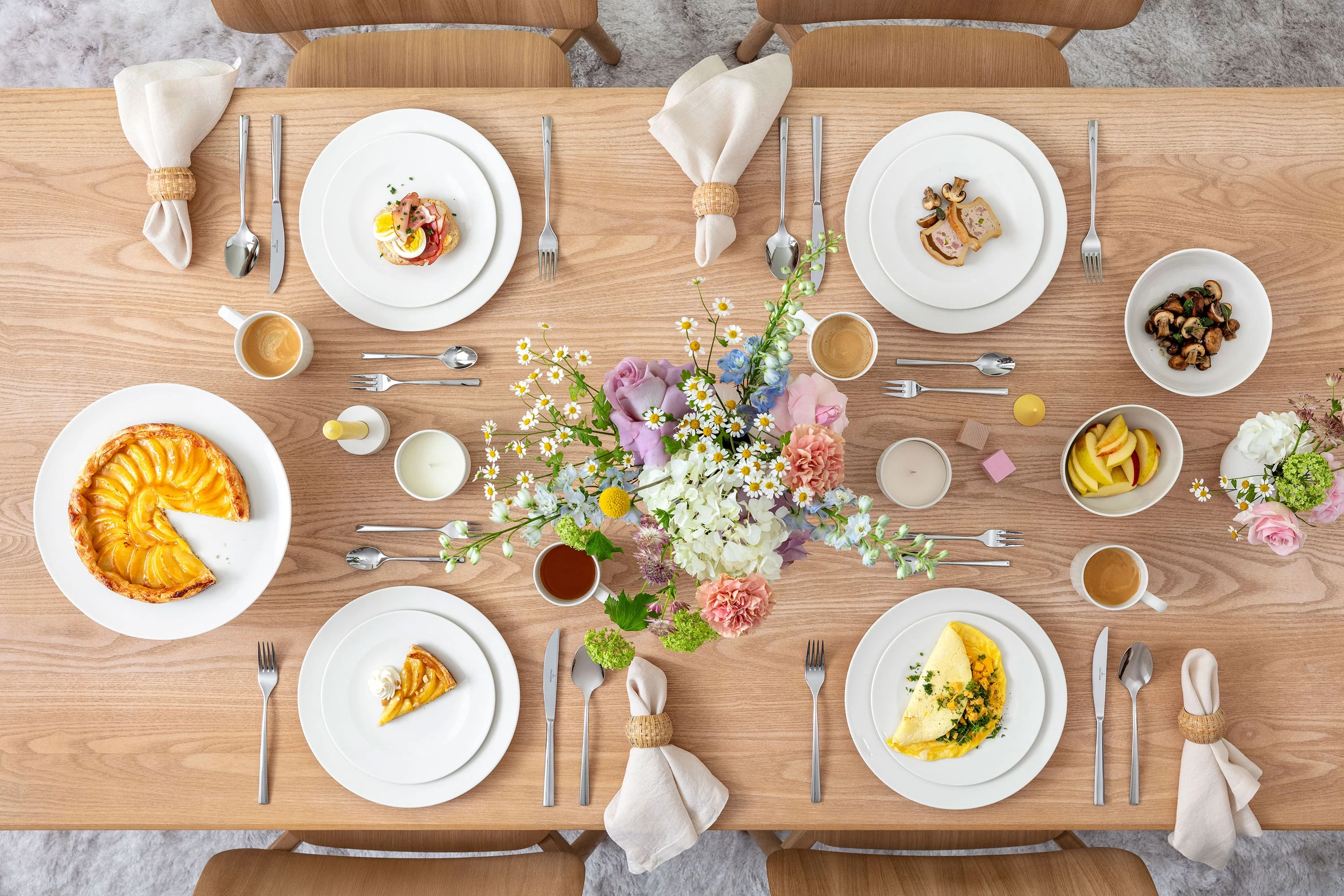 For Me – Inviting Spring Table with Food Draufsicht auf ein frühlingshaft dekoriertes Tischgedeck für vier Personen, angerichtet mit Speisen. Das Arrangement umfasst die For Me Kollektion von Villeroy & Boch: Speiseteller, Frühstücksteller, Schalen, Becher, eine französische Schale und eine Tortenplatte mit Fuß. Jedes Gedeck wird durch das elegante Piemont Besteck ergänzt. Frische saisonale Speisen sorgen für eine lebendige und einladende Atmosphäre. Ein florales Centerpiece mit Frühlingsblumen rundet die Szene auf dem rustikalen Holztisch ab.