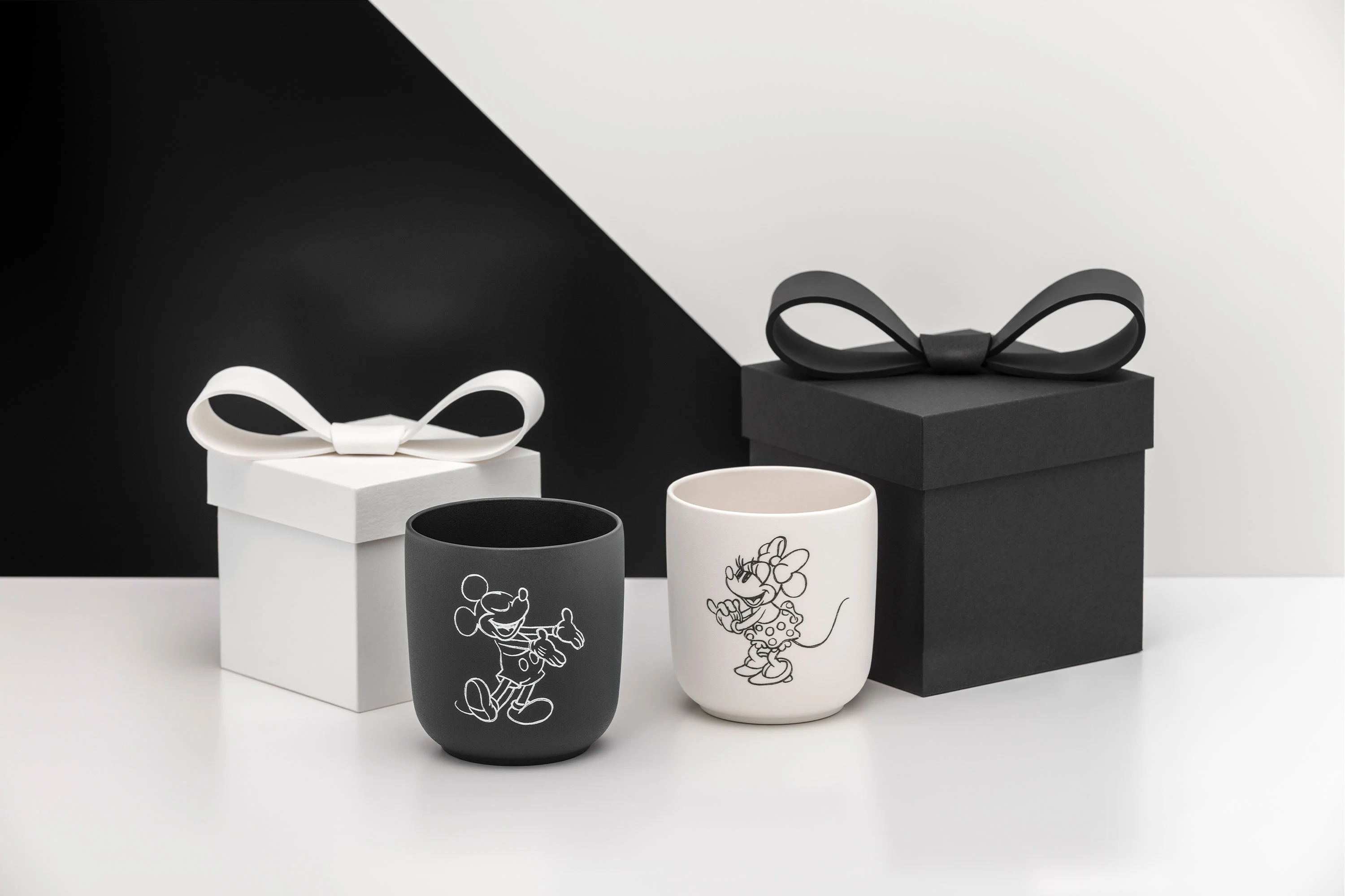 Dwa pudełka upominkowe z kokardkami i dwie pasujące filiżanki Manufacture Mickey, jedna czarna, a druga biała, na białym tle.