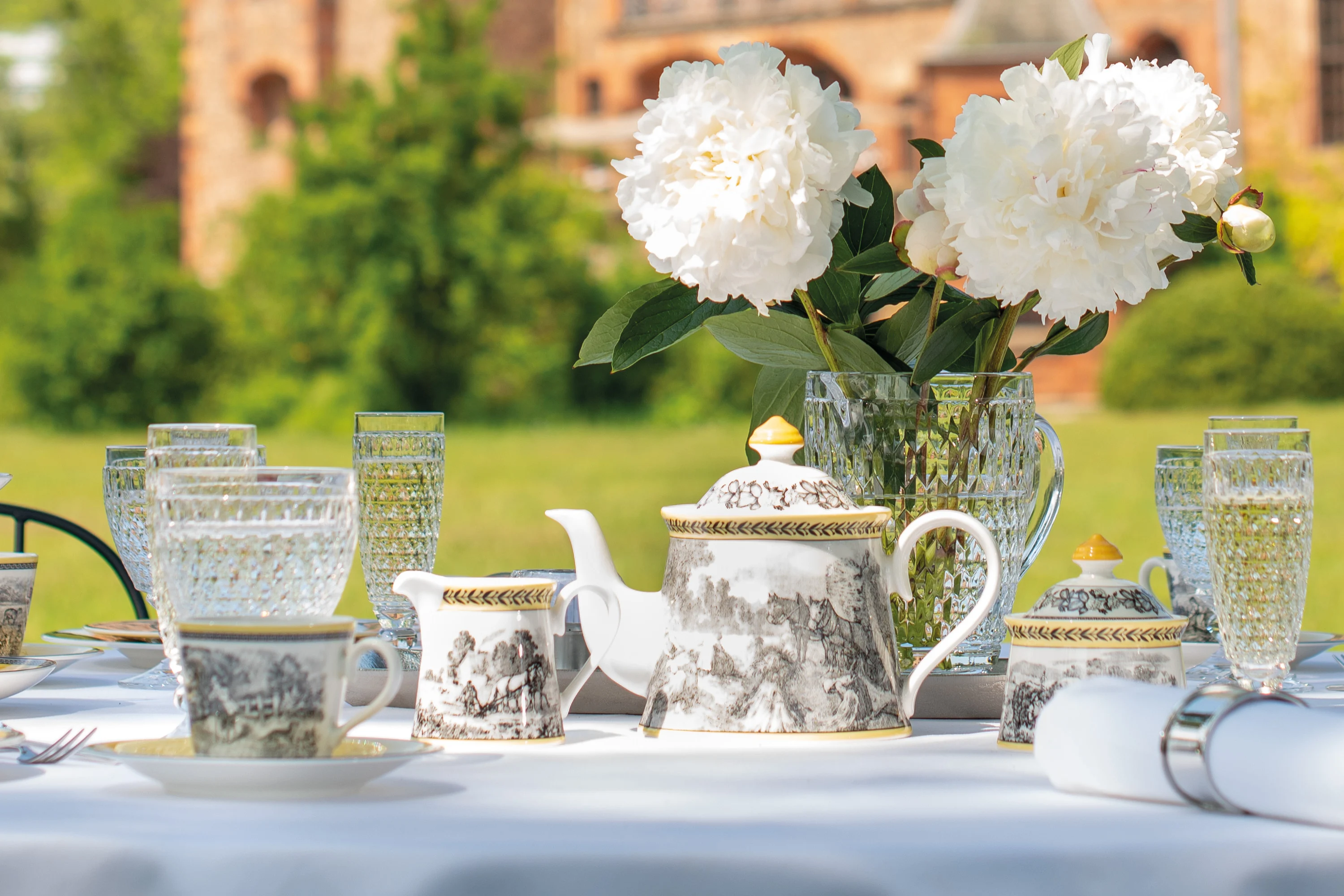 A table set for outdoor tea with ornate teacups and an Audun teapot, crystal glasses and white peonies in a vase. A blurred historic building is visible in the background.