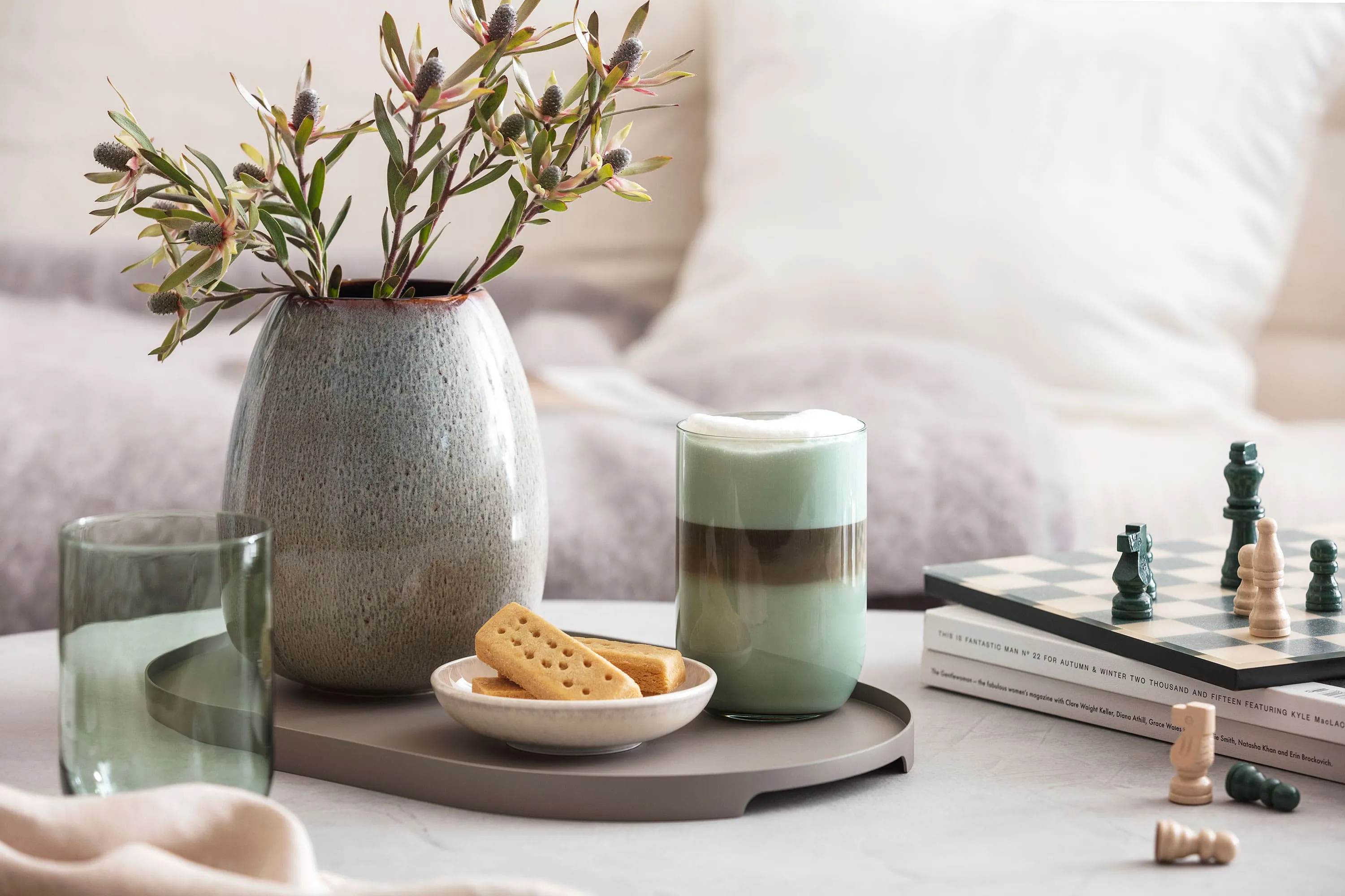Une verre Like Sage avec du café et un vase Lave Home Un verre Like Sage avec du café, un échiquier, des biscuits et un vase Lave avec de l’herbe de la pampa sur un plateau de table.