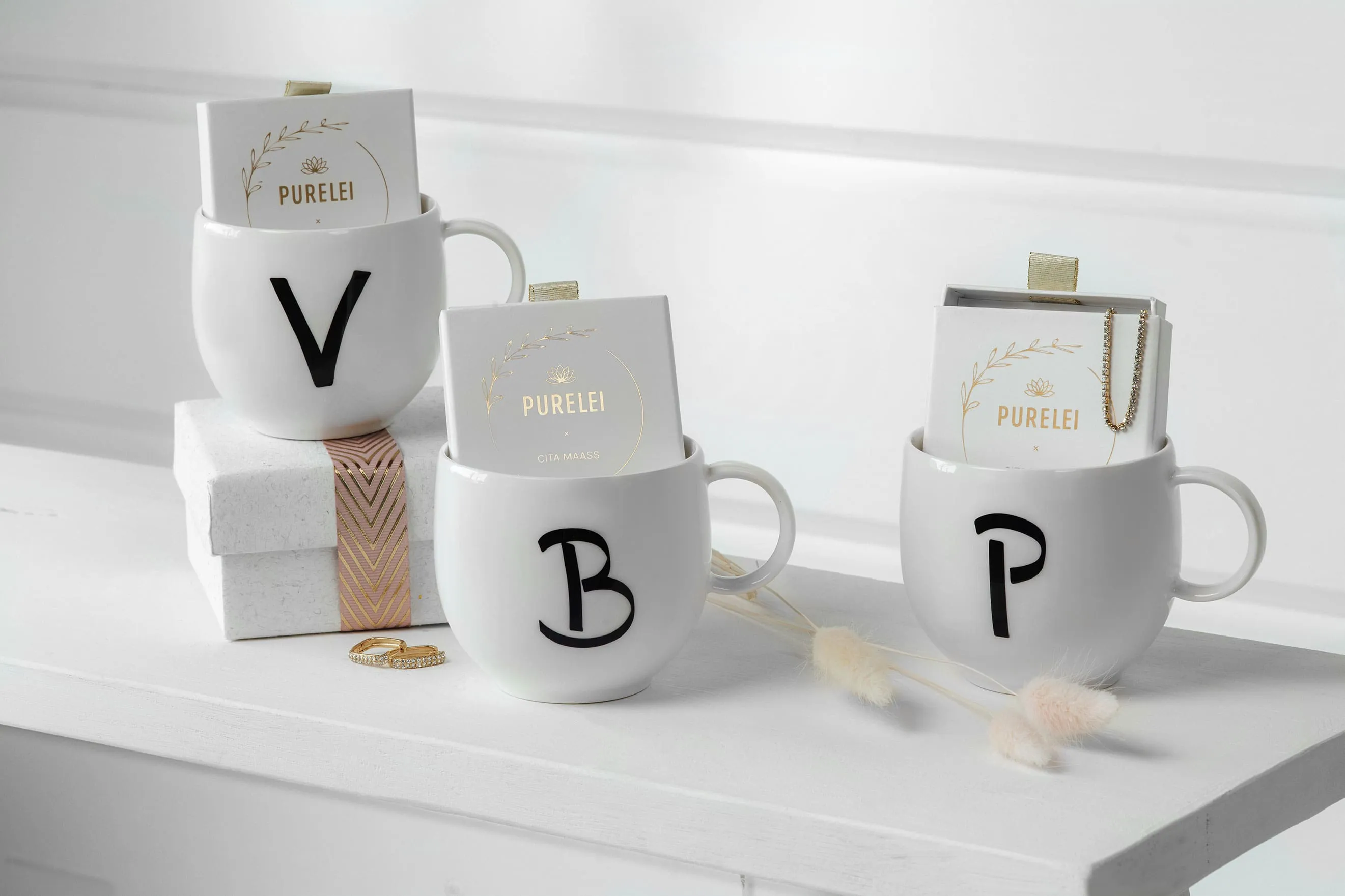 Des mugs Letters portant les lettres V, B et P remplis de bijoux sur une étagère Trois tasses Letters personnalisées portant respectivement les lettres « v », « b » et « p » remplies de bijoux ainsi que des boîtes de produits cosmétiques disposés sur une étagère blanche.