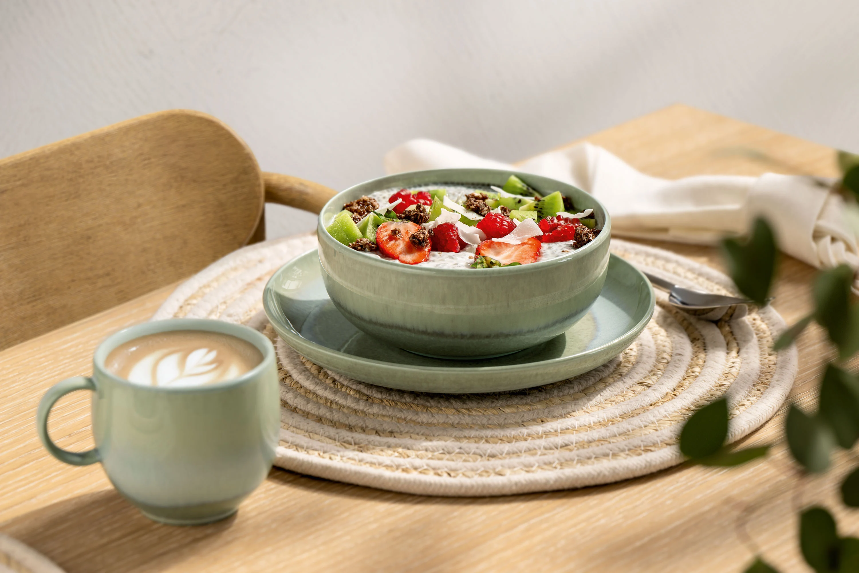 Grüne Perlemor Müsli-Bowl und Kaffe A green Perlemor muesli bowl from Villeroy & Boch with fresh strawberries, kiwis and muesli next to a cup of coffee on a light wooden table.