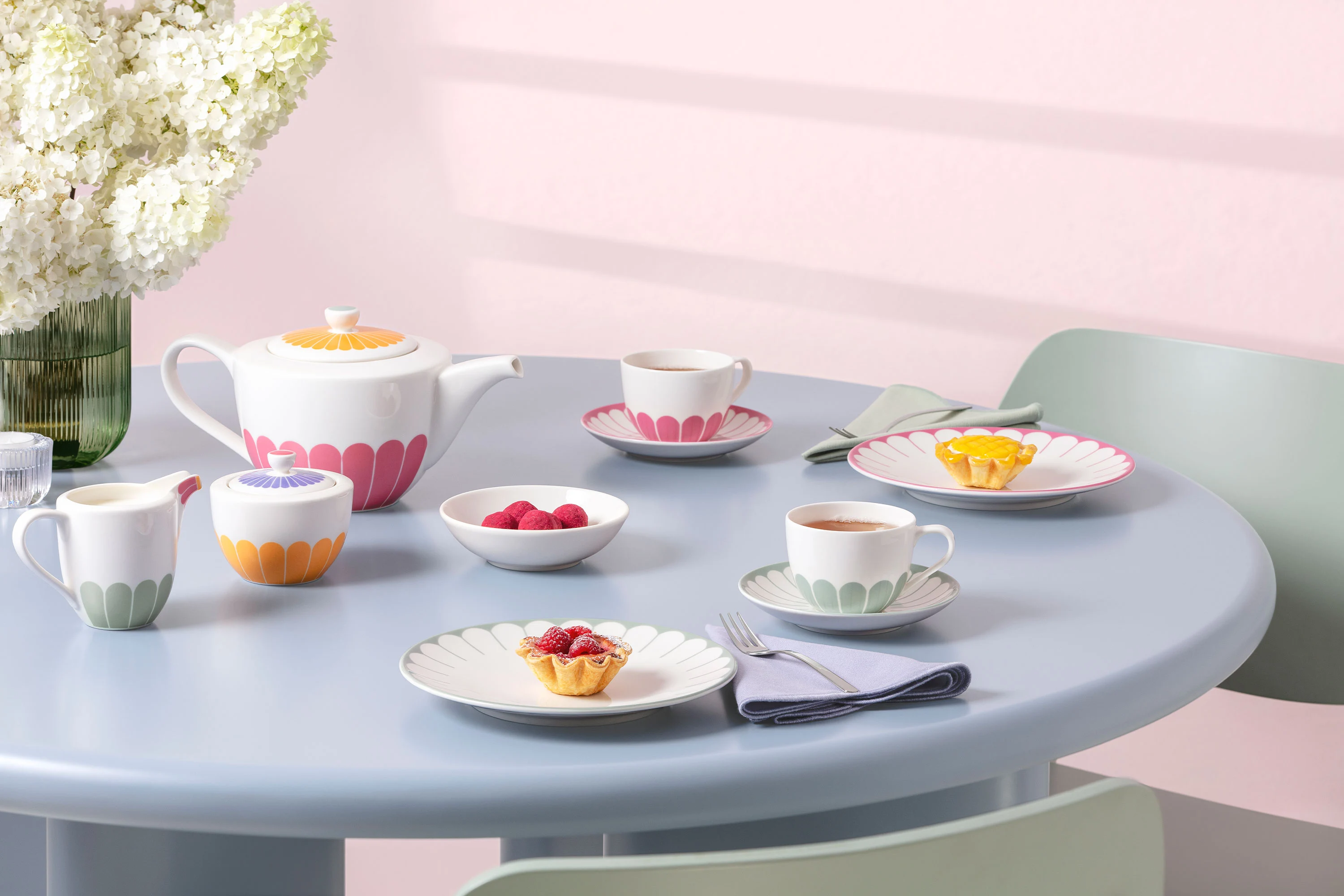 A pastel-coloured tea set by Fleur A pastel-coloured tea set by Fleur with cups, saucers and dessert bowls on a light blue round table next to a vase of white flowers.