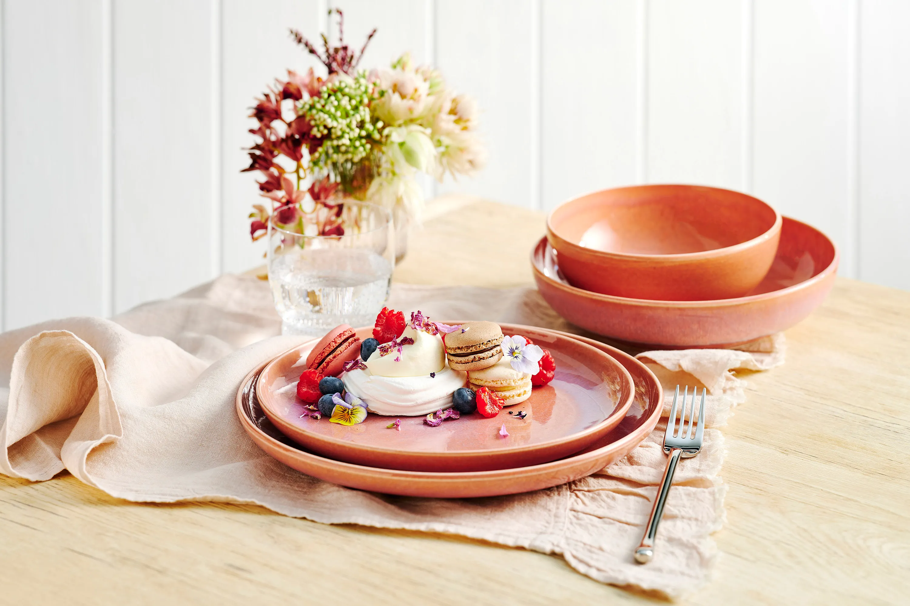 aus-perlemorcoral-indoor-food-nopeople-dinnerset7069 The Perlemor Coral dinner set for one, featuring macarons and fresh fruit, rests on a brown linen tablecloth atop a rustic wooden table, paired with silver cutlery.