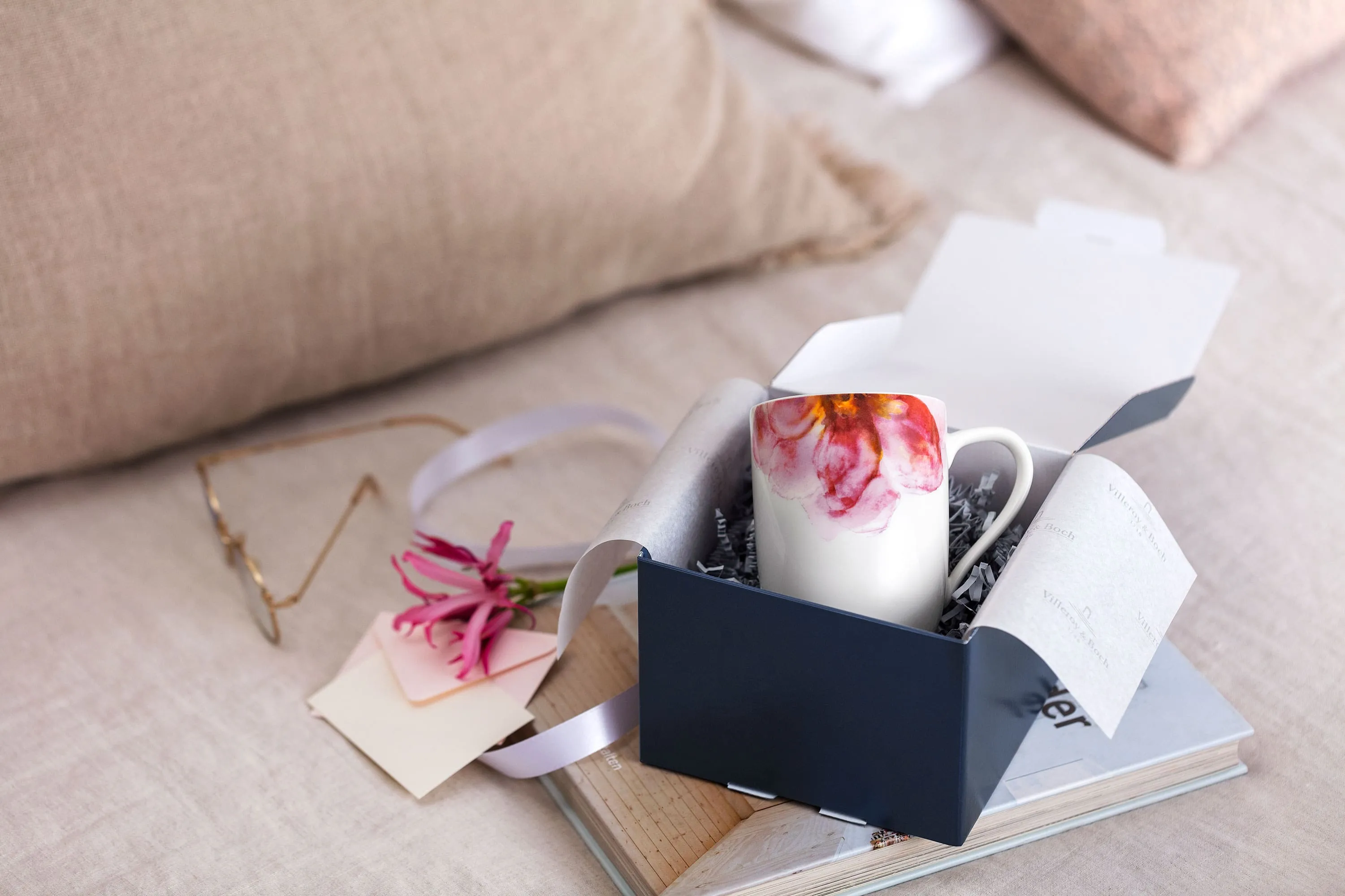 Une tasse Rose Garden dans un emballage cadeau Villeroy & Boch Une boîte cadeau Villeroy & Boch ouverte avec une tasse Rose Garden, des fleurs roses et une note sur un plateau en bois, avec des coussins moelleux en arrière-plan.