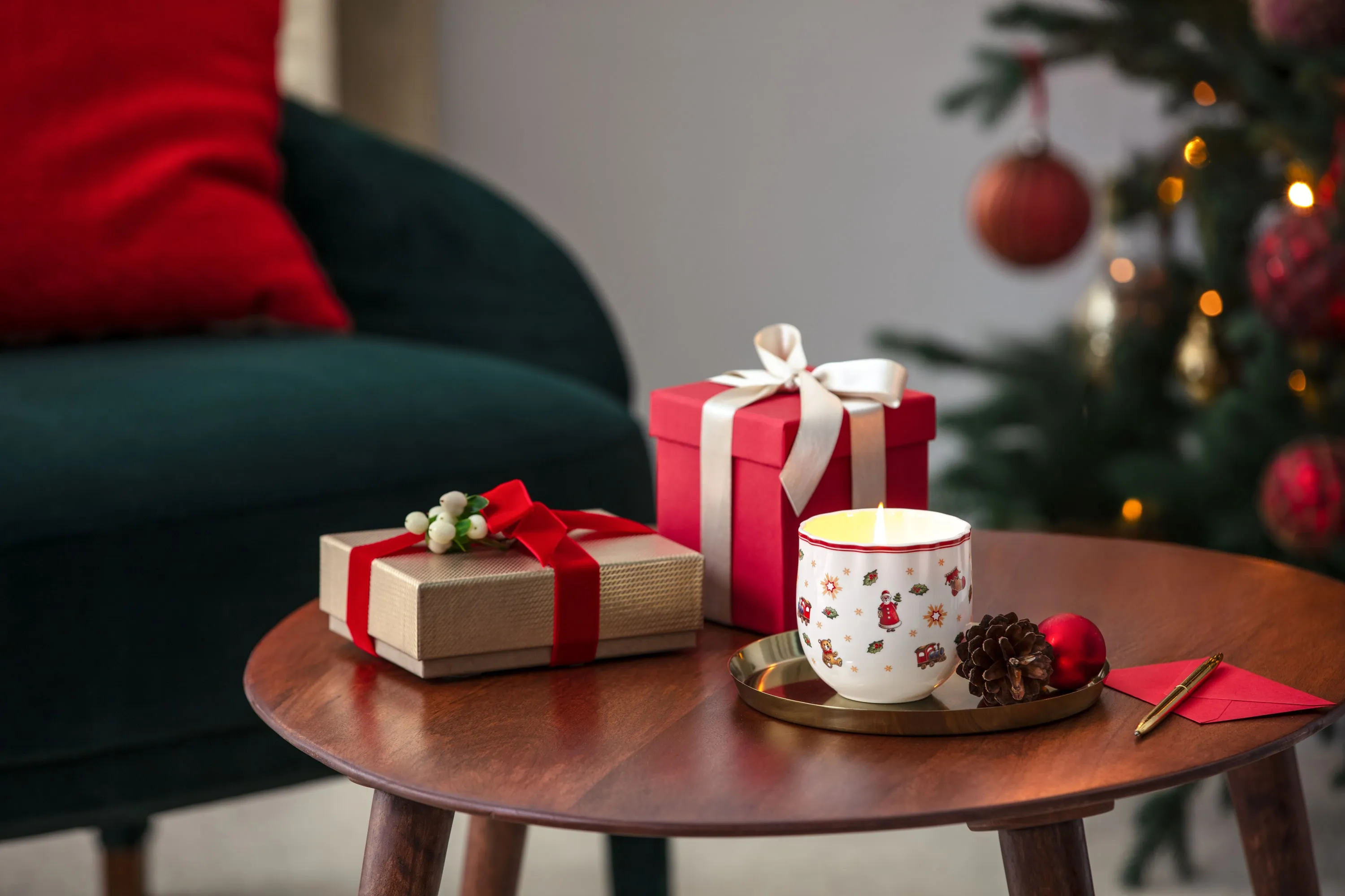 Toy`s Delight Candle as perfect gift A wooden table with two wrapped gifts, a lit candle in a festive mug, pinecones, and a red envelope near a decorated Christmas tree and a green chair.