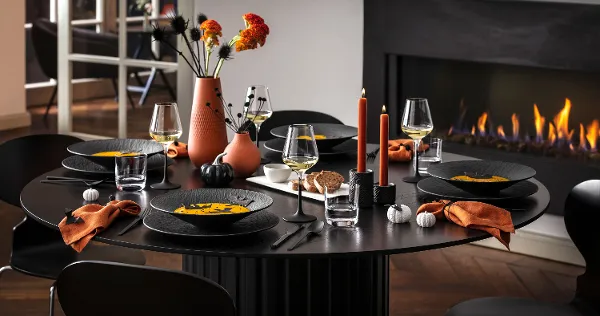 A modern dining table for five with black Manufacture dinnerware, orange napkins, and autumnal decorations, including vases of flowers and lit candles, next to a burning fireplace.