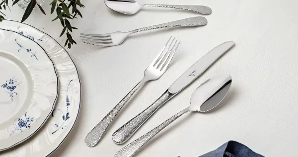Leandra flatware with Vieux Luxembourg plates A Leandra flatware set from Villeroy & Boch is elegantly arranged on a white table. It consists of two forks, two spoons, and a knife. Plates with a blue rim and a blue napkin are partially visible, while green foliage lends the table a decorative charm.