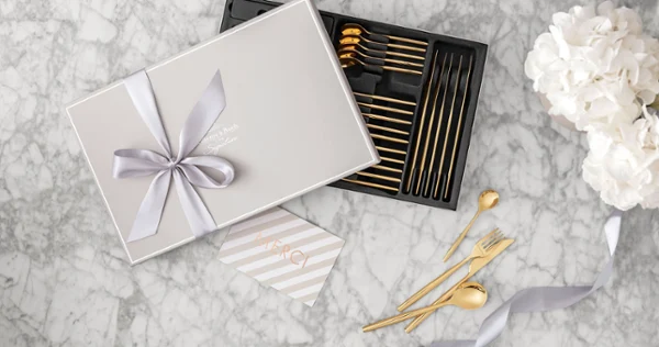 Gift box with golden MetroChic flatware and a bow Gift box with golden MetroChic flatware and a bow, a thank you card and white flowers on a marble surface.
