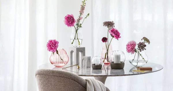 Rose Garden vases and hurricane lamps on a gray marble table A marble table with various flowers in Rose Garden glass vases and Rose Garden hurricane lamps in front of a window with curtains.