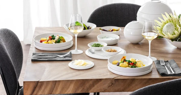 A table set with La Boule with two plates full of food, two glasses of white wine, various bowls with side dishes, and a flower arrangement with black and gray chairs around it.