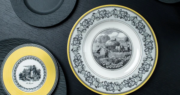 Elegant plates on a dark surface; one large with a rural scene and floral border, another smaller with a yellow rim depicting a couple.