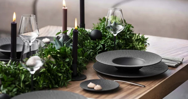 A dining table with black Manufacture plates, flatware, wine glasses, and dark candles. Green plants are used as table decorations and create a sophisticated atmosphere.