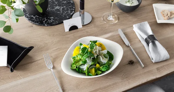 A Vapiano bowl from Villeroy & Boch with salad on a wooden table, next to it a knife and fork from NewWave.