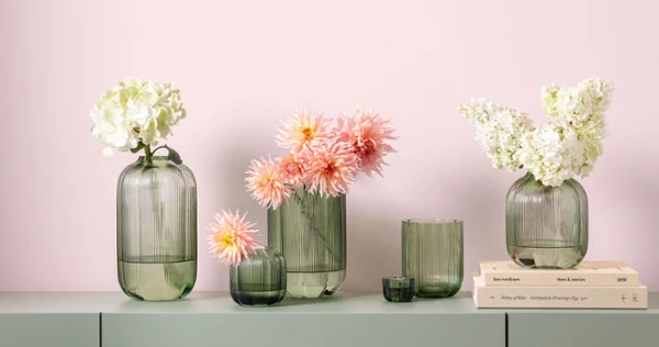 Green glass vases from Fleur with white and pink flowers arranged on a light green surface in front of a light pink wall.