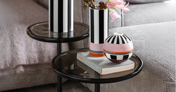 Two black and white striped vases and a round vessel stand on glass tables next to a grey sofa, flowers in a vase