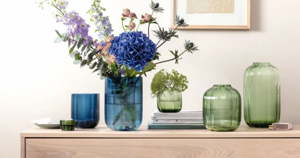 Green and blue glass vases in various shapes some with flowers in them standing on wood furniture with a picture on the wall behind them