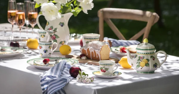 Outdoor table setting with flowered French Garden china, a teapot, rosé glasses, and a cake. White peonies in a vase and fruit are placed around the table.