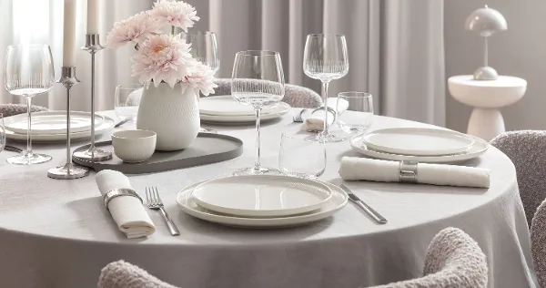 Table de salle à manger ronde avec des assiettes Afina blanches, des verres Afina, des couverts, des serviettes et un vase avec des fleurs roses.