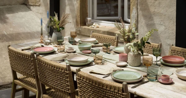 A rustic outdoor dining table set with woven chairs, pastel-colored Perlemor plates from like. by Villeroy & Boch, glasses and floral centerpieces against the backdrop of a stone wall with a window.