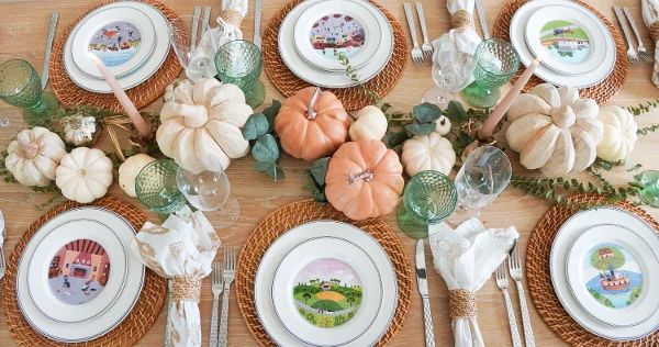 A wooden dining table set for a meal with decorative pumpkins, green glasses, white plates with illustrated scenes, and natural woven placemats.