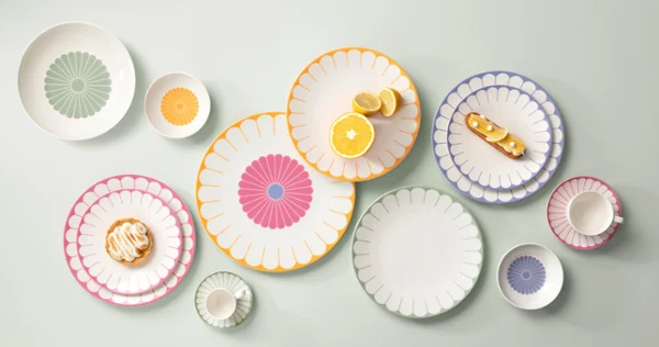 Various colourful, patterned plates, bowls and a cup are arranged on a light-coloured surface, each with a different design. Pastries are placed on some of the plates.	