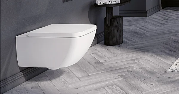 Toaleta w kolorze białym z kolekcji Collaro Nowoczesna łazienka z wiszącą białą toaletą Villeroy & Boch, ciemną półką z dekoracjami i małym stolikiem z rośliną. Pomieszczenie ma szare ściany i podłogę ze wzorem w jodełkę.