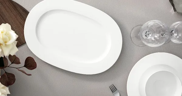 A table setting with a large white oval platter, a white plate, a bowl, a fork, wine glasses and decorative white flowers on a textured grey tablecloth. Everything from Royal by Villeroy & Boch.