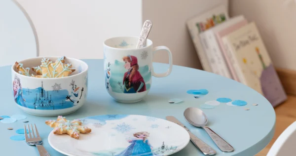 A children's table with chair and Frozen-style tableware, cereal, cookies in the background.