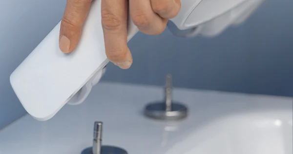 Close-up of a hand holding a bidet nozzle over a white bathroom fitting with two control elements against a blue background.