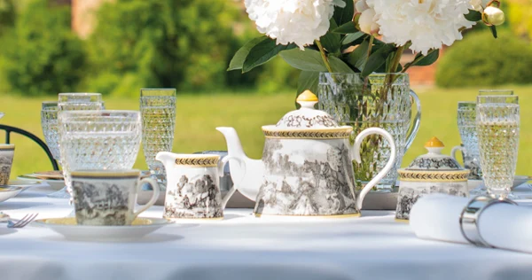 Outdoor tea set with Audun dinnerware A table set for outdoor tea with ornate teacups and an Audun teapot, crystal glasses, and white peonies in a vase. A historic building is blurred in the background.