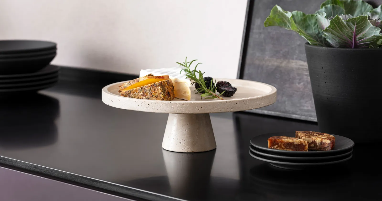Perfect serving platter in Travertine for antipasti or cheese. A selection of cheese on a Travertine serving platter with a foot, and dark fruits, next to black Manufacture tableware by Villeroy & Boch on a dark worktop.