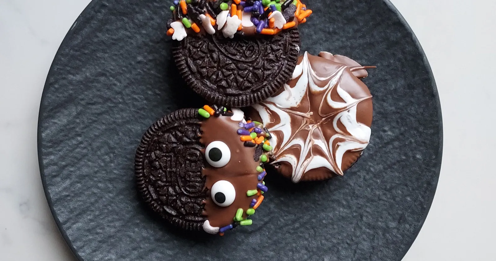 Three Halloween cookies on a black plate: one with candy eyes and sprinkles, another with a web design.