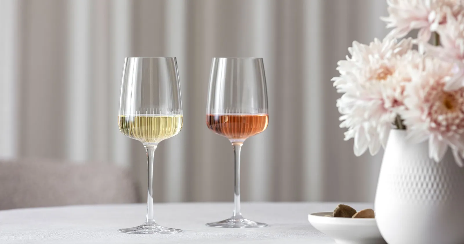 Two Villeroy & Boch Afina wine glasses, one filled with white wine and the other with rosé, are placed on a table next to a vase with pink flowers.