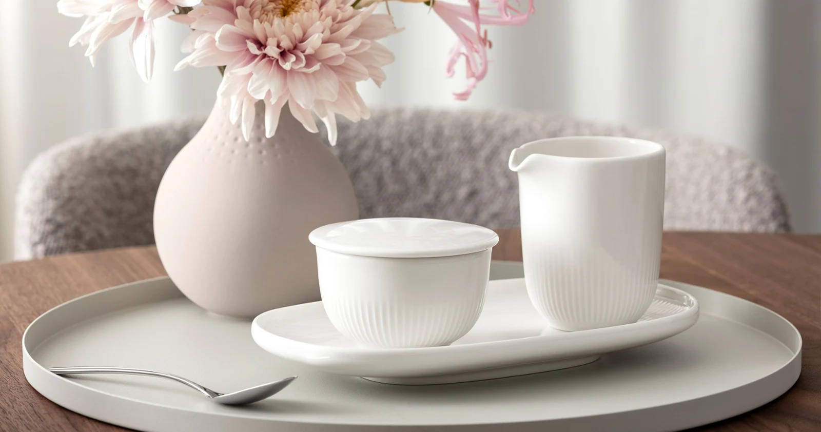 An Afina milk jug and an Afina sugar bowl sit on an oval tray An Afina milk jug and an Afina sugar bowl sit on an oval tray from the same collection, in front of a Manufacture vase containing pink flowers.